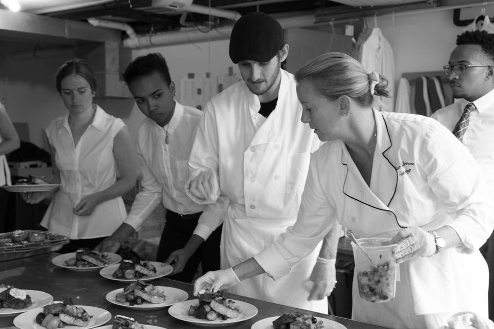 Five culinary professionals in white chef coats standing in a kitchen, gathering around a counter with plated dishes. A chef in a black cap demonstrating plating techniques while others observe, with one person holding a clear container of ingredients.